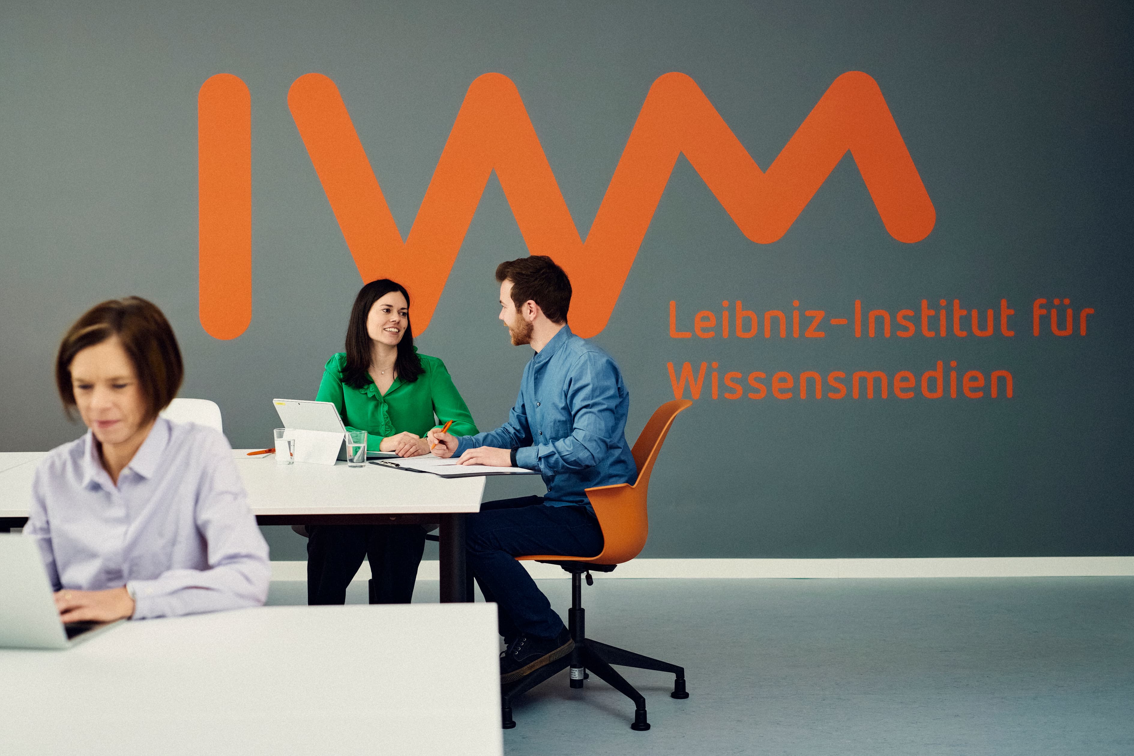 Drei IWM-Mitarbeitende arbeiten vor einem großen IWM-Schriftzug im Hintergrund